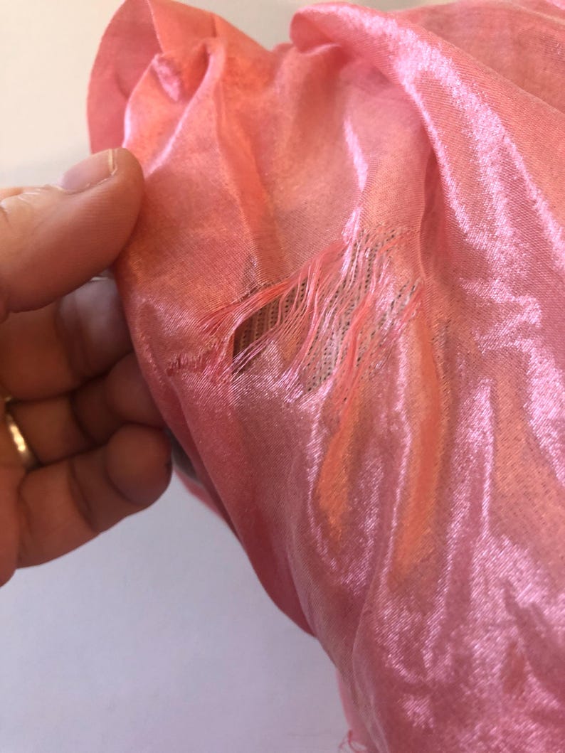 May include: Close-up of a section of a pink fabric, possibly silk or a similar material. The fabric has a shiny, reflective quality. There is a tear in the fabric, with frayed edges visible. The fabric is being held by a hand.