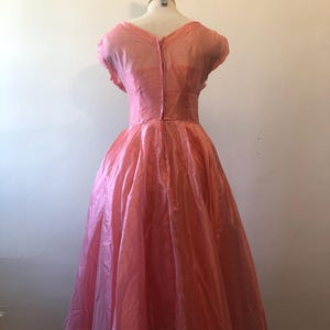 May include: A vintage pink dress displayed on a mannequin. The dress features a fitted bodice, a V-neckline, and a full skirt. The fabric has a slight sheen, and the dress appears to be knee-length. The dress is a soft, light pink color.