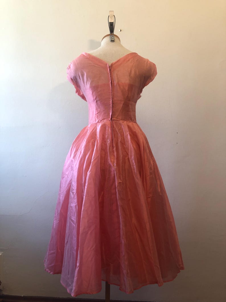 May include: A vintage pink dress displayed on a mannequin. The dress features a fitted bodice, a V-neckline, and a full skirt. The fabric has a slight sheen, and the dress appears to be knee-length. The dress is a soft, light pink color.