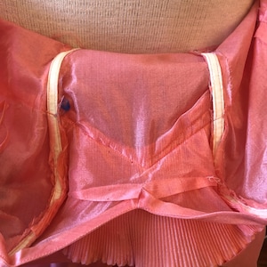 May include: Close-up of a pink, sheer garment with a pleated bottom edge. The garment has a V-shaped design at the waist and white trim along the edges. The fabric appears to be lightweight and flowing.