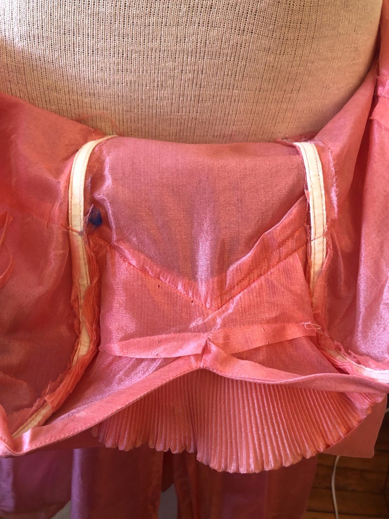 May include: Close-up of a pink, sheer garment with a pleated bottom edge. The garment has a V-shaped design at the waist and white trim along the edges. The fabric appears to be lightweight and flowing.