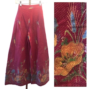 May include: Wide-leg, high-waisted pants in a deep red hue, adorned with embroidered floral and wheat designs in shades of yellow, blue, and green. The pants feature a detailed close-up of the embroidery, showcasing the intricate craftsmanship and texture of the fabric.