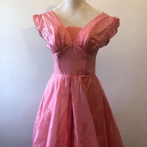 May include: A vintage pink dress with a V-neckline and short, puffed sleeves. The dress features a fitted bodice and a full, pleated skirt. The fabric has a subtle sheen, giving it a formal appearance. The dress is displayed on a mannequin.