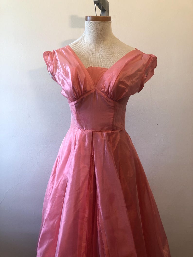May include: A vintage pink dress with a V-neckline and short, puffed sleeves. The dress features a fitted bodice and a full, pleated skirt. The fabric has a subtle sheen, giving it a formal appearance. The dress is displayed on a mannequin.