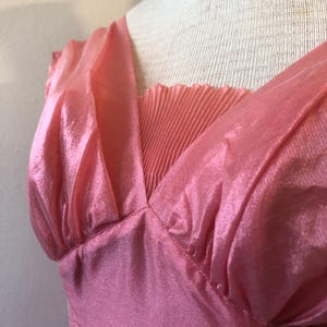 May include: A pink dress with a V-neckline and pleated detail. The dress is made of a shiny, flowing fabric and has a gathered shoulder detail. The dress is displayed on a mannequin.