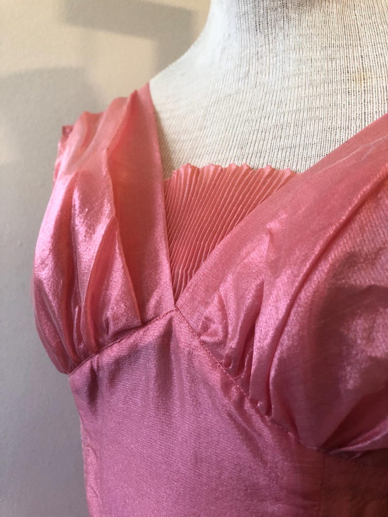 May include: A pink dress with a V-neckline and pleated detail. The dress is made of a shiny, flowing fabric and has a gathered shoulder detail. The dress is displayed on a mannequin.