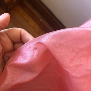 May include: Close-up of a pink, translucent fabric. The fabric has a slight sheen and appears to be thin and delicate. A small, dark mark is visible on the fabric's surface. The fabric is likely used for clothing or decorative purposes.