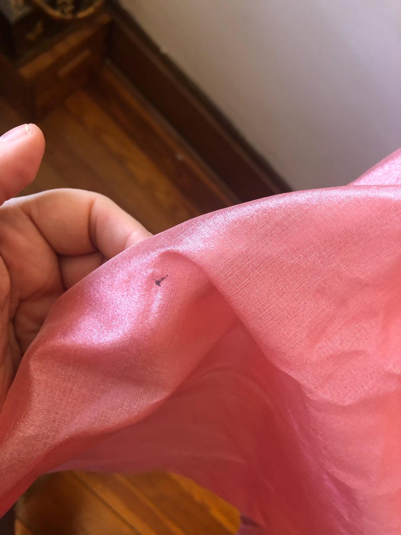 May include: Close-up of a pink, translucent fabric. The fabric has a slight sheen and appears to be thin and delicate. A small, dark mark is visible on the fabric's surface. The fabric is likely used for clothing or decorative purposes.