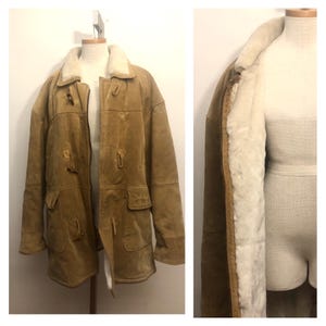 May include: A tan suede coat with a cream-colored shearling lining and collar. The coat features toggle closures, two front pockets, and is displayed on a mannequin. The coat's interior is visible, showcasing the warm shearling.