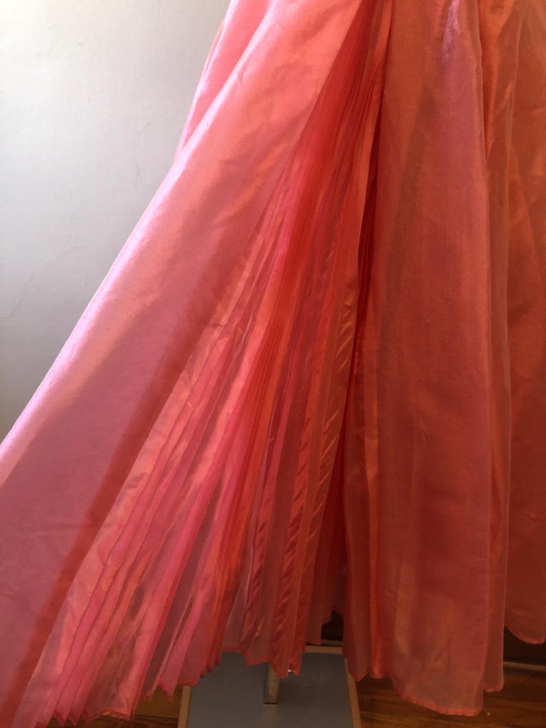 May include: A flowing, coral-colored garment with a pleated section. The fabric appears to be lightweight and has a slight sheen, suggesting a satin or silk-like material. The garment is likely a dress or skirt.