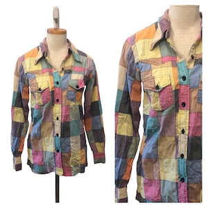 May include: A long-sleeve button-down shirt featuring a multi-colored patchwork pattern in shades of yellow, pink, blue, orange, and tan. The shirt includes a pointed collar, two chest pockets with button flaps, and dark buttons down the front.