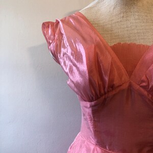 May include: A vintage pink dress with a V-neckline and pleated bodice. The dress features short, ruffled sleeves and a flowing skirt. The fabric has a shiny, iridescent quality, giving it a glamorous appearance.