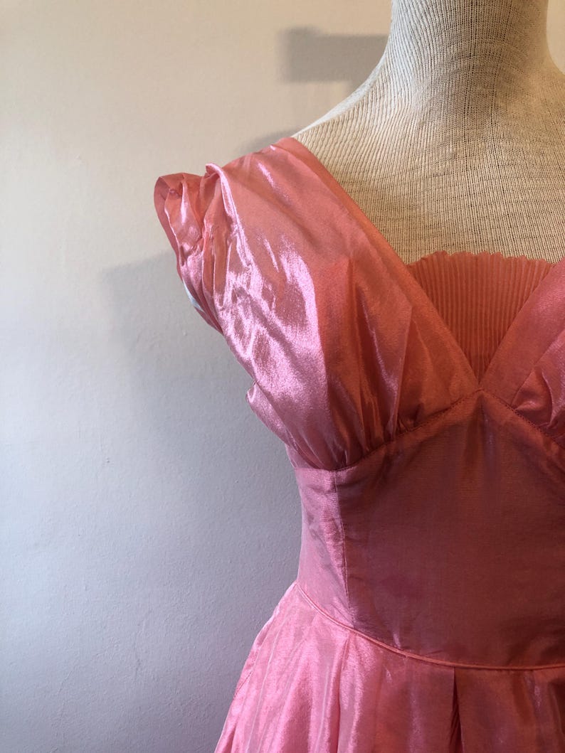 May include: A vintage pink dress with a V-neckline and pleated bodice. The dress features short, ruffled sleeves and a flowing skirt. The fabric has a shiny, iridescent quality, giving it a glamorous appearance.