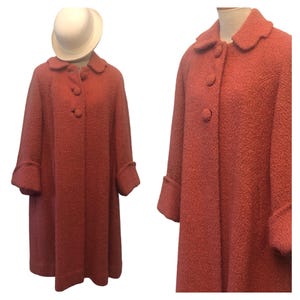 May include: A textured coral-colored coat featuring a rounded collar, three large matching fabric-covered buttons down the front, and cuffed sleeves. The coat has a straight, structured silhouette and is displayed alongside a white bowler hat.