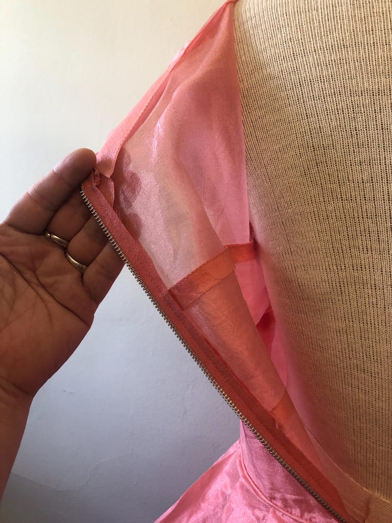 May include: Close-up of a pink dress with a zipper closure. The dress is made of a shiny, silky material. The zipper is silver and runs along the side of the dress. The dress is displayed on a mannequin.