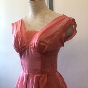 May include: A vintage coral-colored dress displayed on a mannequin. The dress features a V-neckline, pleated bodice, and a full skirt. The fabric appears to be a lightweight, possibly silk or chiffon, with short, puffed sleeves. The dress is a classic style.