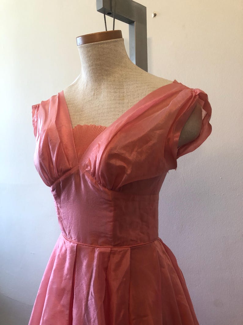 May include: A vintage coral-colored dress displayed on a mannequin. The dress features a V-neckline, pleated bodice, and a full skirt. The fabric appears to be a lightweight, possibly silk or chiffon, with short, puffed sleeves. The dress is a classic style.