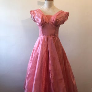 May include: A vintage pink dress with a V-neckline and short, puffed sleeves. The dress features a fitted bodice and a full, flowing skirt. The fabric has a subtle sheen, and the dress is displayed on a mannequin.