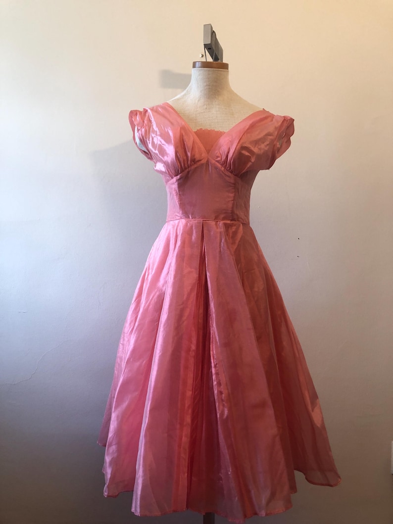 May include: A vintage pink dress with a V-neckline and short, puffed sleeves. The dress features a fitted bodice and a full, flowing skirt. The fabric has a subtle sheen, and the dress is displayed on a mannequin.