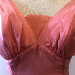 May include: A vintage-style, coral-pink dress with a V-neckline. The bodice features a pleated insert and gathered fabric at the shoulders. The dress is displayed on a mannequin, showcasing its elegant design and flowing silhouette.