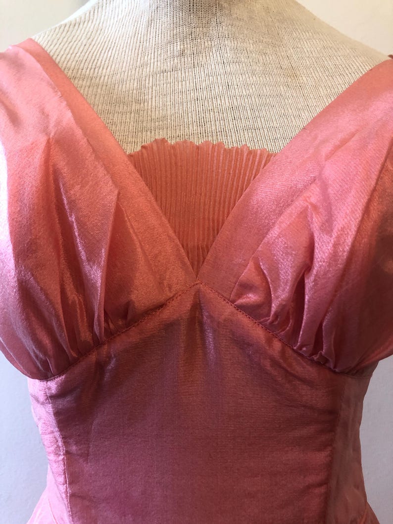 May include: A vintage-style, coral-pink dress with a V-neckline. The bodice features a pleated insert and gathered fabric at the shoulders. The dress is displayed on a mannequin, showcasing its elegant design and flowing silhouette.