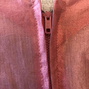 May include: Close-up of a pink garment with a zipper. The fabric appears sheer and has a subtle sheen. The zipper is a matching pink color, and the stitching is visible along the seams.