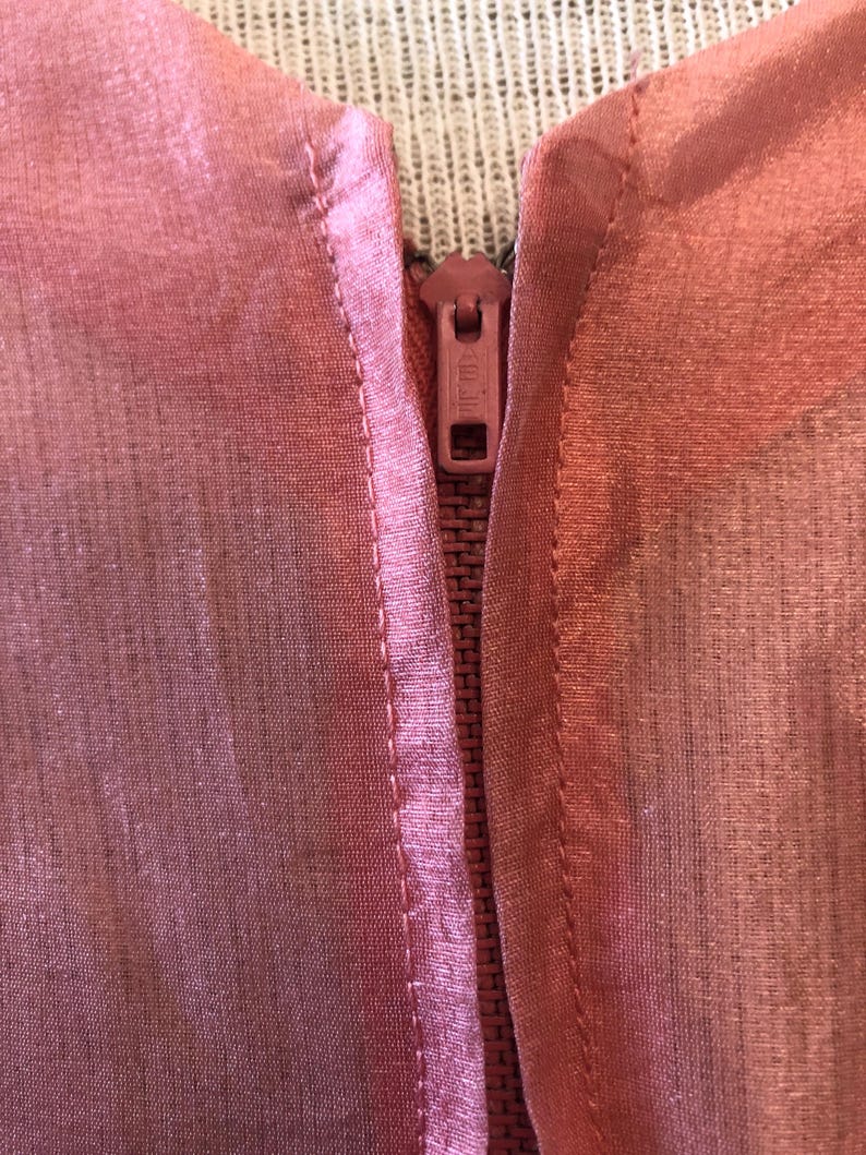 May include: Close-up of a pink garment with a zipper. The fabric appears sheer and has a subtle sheen. The zipper is a matching pink color, and the stitching is visible along the seams.