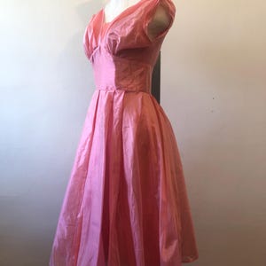 May include: A vintage pink dress displayed on a mannequin. The dress features a fitted bodice with a V-neckline and short, puffed sleeves. The skirt is full and flowing, creating a classic silhouette. The fabric has a subtle sheen, suggesting a silk or satin material.
