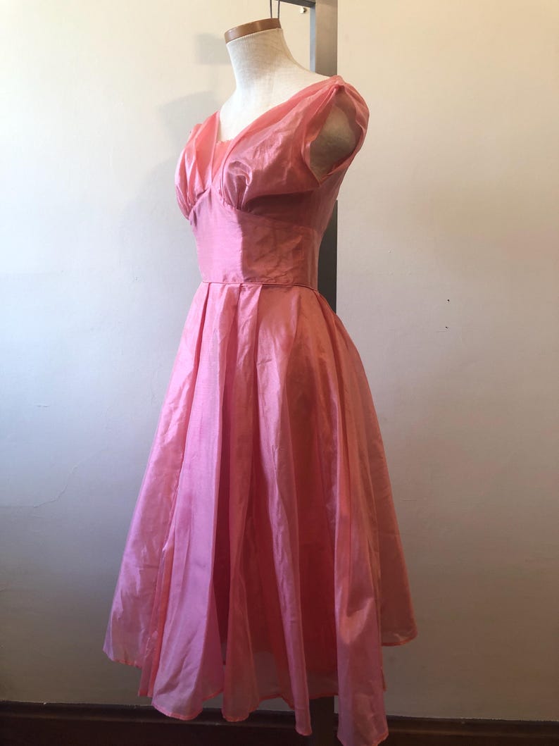 May include: A vintage pink dress displayed on a mannequin. The dress features a fitted bodice with a V-neckline and short, puffed sleeves. The skirt is full and flowing, creating a classic silhouette. The fabric has a subtle sheen, suggesting a silk or satin material.