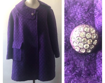 May include: A deep purple coat with a textured diamond pattern, featuring a wide collar and a single large button embellished with multiple clear, faceted crystals. The coat has long sleeves and two front pockets with flaps.