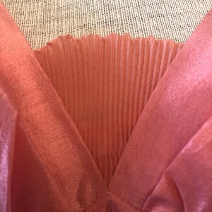 May include: Close-up of a coral-colored garment with a V-neck design. The fabric has a silky, flowing texture, with a pleated panel at the neckline. The garment appears to be a top or dress, showcasing a delicate and feminine style.