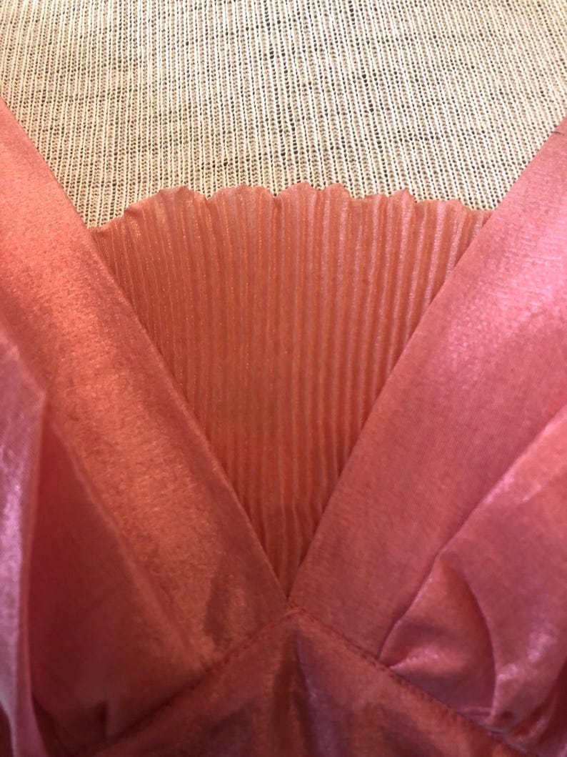 May include: Close-up of a coral-colored garment with a V-neck design. The fabric has a silky, flowing texture, with a pleated panel at the neckline. The garment appears to be a top or dress, showcasing a delicate and feminine style.