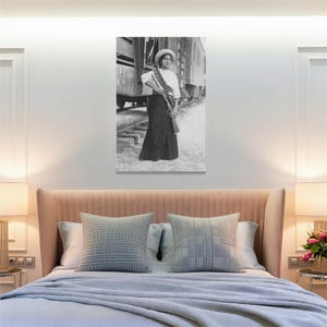 May include: Black and white print of a woman in a long skirt and hat, holding a rifle. She stands near a train car on railroad tracks. The image is a vintage photograph, likely from the early 20th century.