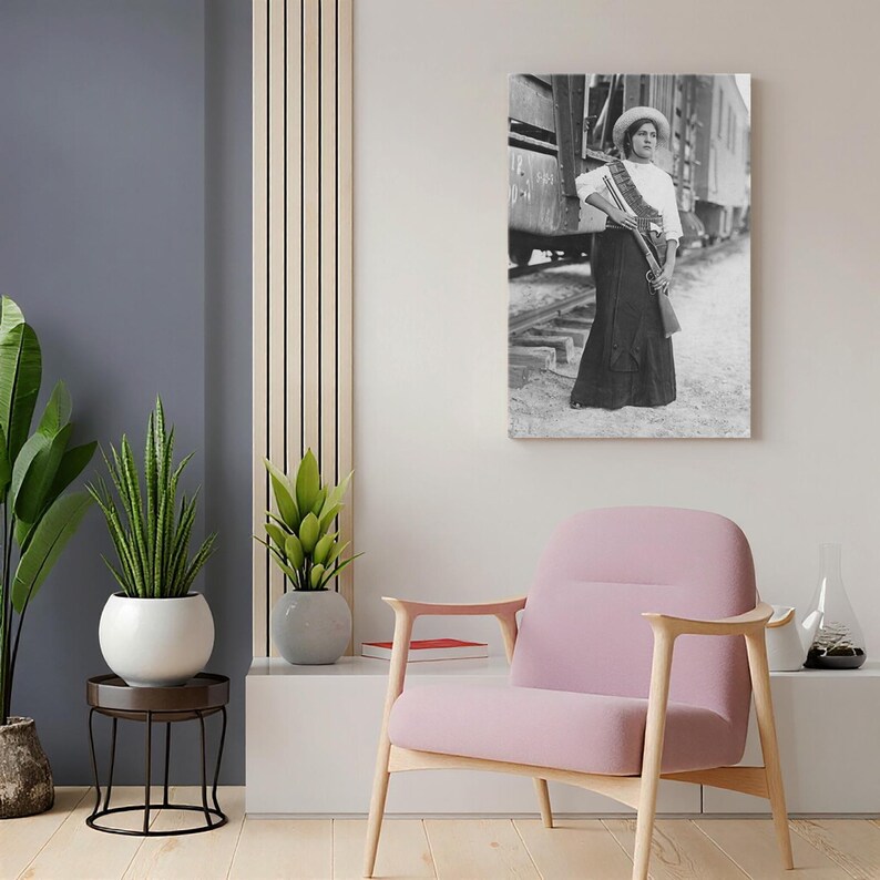 May include: Black and white print of a woman in a long skirt, white blouse, and hat, holding a rifle. She stands near a train, with a sash across her chest. The image is mounted on a wall, part of a room's decor.