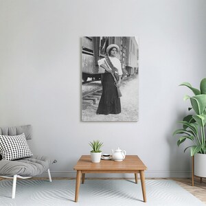 May include: Black and white print of a woman in a long skirt, white blouse, and hat, holding a rifle. She stands near a train, with a bandolier across her chest. The image is a vintage photograph, likely from the early 20th century.