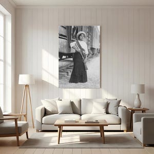 May include: Black and white print of a woman in a long skirt, white blouse, and hat, holding a rifle. She stands near a train, wearing a sash. The image is a vintage photograph, likely from the early 20th century, and is displayed as wall art.