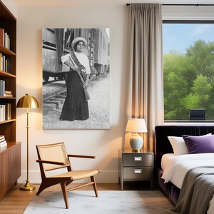 May include: Black and white framed print of a woman in a white blouse, long skirt, and hat, holding a rifle and standing near a train. The print is displayed in a modern bedroom with a gold floor lamp, wooden chair, and a window with a view of trees.