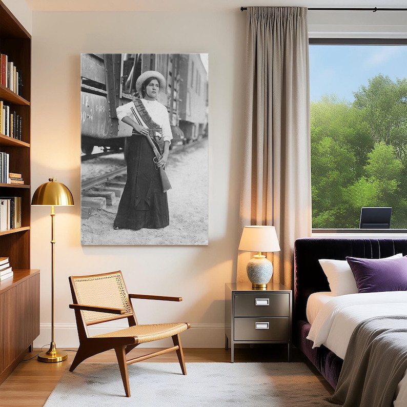May include: Black and white framed print of a woman in a white blouse, long skirt, and hat, holding a rifle and standing near a train. The print is displayed in a modern bedroom with a gold floor lamp, wooden chair, and a window with a view of trees.