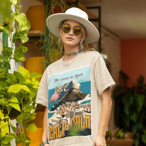 May include: Beige t-shirt featuring a colorful graphic of a flamingo, mountain, and city with the text "The Peñón de Ifach" and "Calp & Calm". The shirt is paired with a white hat and sunglasses.