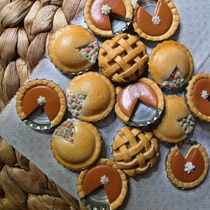 May include: A collection of miniature pie-shaped ornaments crafted from bottle caps. The set includes smooth brown pumpkin-style pies with white dollops and textured savory pies filled with colorful green, orange, and white vegetable pieces.