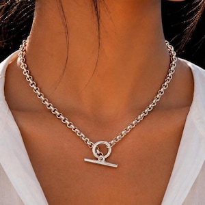 May include: A silver-toned chain necklace featuring a circular ring and a horizontal bar toggle clasp closure. The necklace is constructed from uniform round links that form a continuous chain around the neck, meeting at the front center.