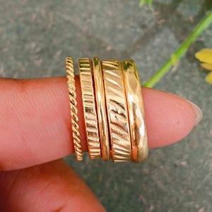 May include: A set of five gold-toned rings worn on a finger. The collection includes one thin twisted rope band, two flat bands with vertical etched lines, one smooth thin band, and one wider band with a hammered, uneven surface texture.
