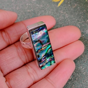 May include: A rectangular silver ring featuring a vibrant abalone shell inlay. The iridescent shell displays a mix of blues, greens, and purples. The ring's band is a simple, polished silver design.