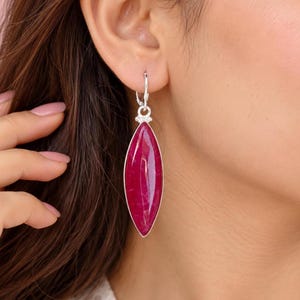 Può includere: Un orecchino lungo a forma di marquise con una gemma liscia color magenta intenso, incastonata in una montatura in metallo color argento lucido. L'orecchino pende da una semplice chiusura a monachella color argento.