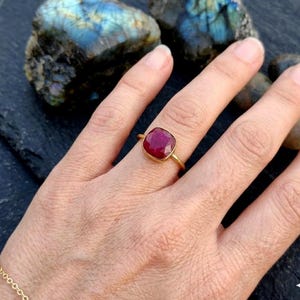 May include: A gold ring featuring a square-cut, faceted deep red gemstone set in a simple metal band. The ring is worn on a finger, showcasing the vibrant color and geometric shape of the stone against a dark, textured background.