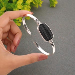May include: A silver cuff bracelet featuring a central rectangular black tourmaline stone. The stone has a vertically striated texture and is set within a polished silver frame. The band is open-ended, allowing for adjustment on the wrist.