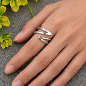 May include: A silver-toned ring featuring an open, bypass-style design with two pointed, angular ends that overlap slightly. The metal has a smooth, polished finish, creating a clean and reflective surface that wraps around the finger.