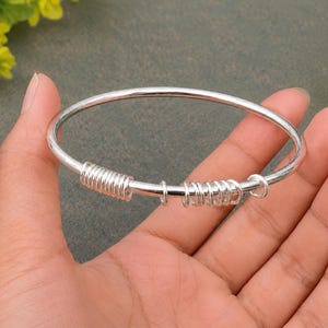 May include: A silver-colored bangle bracelet featuring a smooth, circular band adorned with two distinct sections of coiled wire detailing and several small, loose silver-colored rings positioned along the circumference of the metal band.