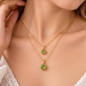 May include: A layered gold necklace set featuring two delicate chains, each adorned with a single round, bezel-set peridot gemstone pendant. The pendants are circular with a polished gold finish, showcasing the vibrant green colour of the stones.