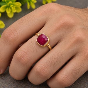 Peut inclure: Une bague en or ornée d'une pierre précieuse rouge foncé taillée en coussin à facettes, sertie dans une monture en métal lisse. L'anneau est fin et poli, mettant en valeur la couleur vive et la forme géométrique de la pierre centrale.